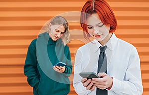 Smiling caucasian teenage girl portrait with red dyed hair and friend boy behind browsing their smartphone devices. Careless young