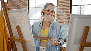 A smiling caucasian woman artist in a denim shirt holds a palette in a bright art studio photo