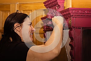 Smiling Caucasian woman coloring wooden ornamented cupboard in red with large paint brush with doors in background