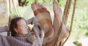 Smiling caucasian woman lying barefoot in hammock, relaxing with eyes closed in sunny garden