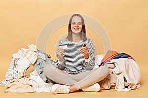 Smiling Caucasian woman posing near heap of multicolored unsorted clothes isolated over beige background buying attires online