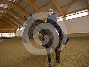 Smiling Caucasian woman sitting on beautiful brown horse while riding it indoors