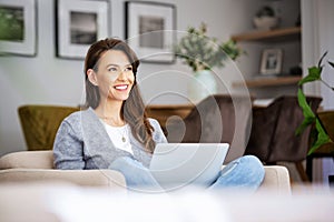 Smiling caucasian woman using laptop at home while sitting in an armchair