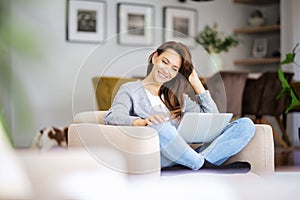 Smiling caucasian woman using laptop at home while sitting in an armchair