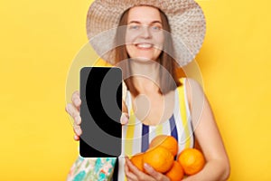 Smiling caucasian woman wearing striped swimsuit and straw hat isolated yellow background showing empty smart phone screen