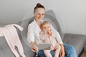 Smiling Caucasian woman wearing white shirt and jeans sitting on sofa with baby daughter, holding mobile phone and taking selfie