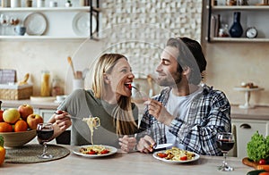 Smiling caucasian young male with stubble feeds blonde woman, couple have fun together in light kitchen interior