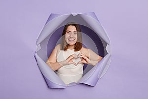 Smiling cheerful brunette woman breaking through purple paper hole wearing casual white top showing heart gesture with hands
