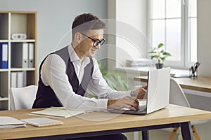 Smiling cheerful business man working on a laptop at workplace and typing in office.