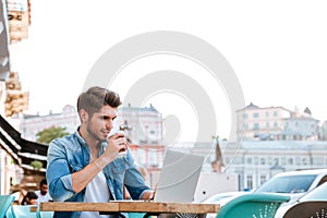 Smiling cheerful casual man using laptop outdoors and drinking coffee