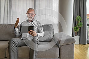 Smiling cheerful happy senior man waving and gesturing to camera having online video call on hand held tablet portable device