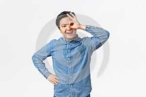 Smiling cheerful and satisfied asian man showing okay gesture over eye, looking through with pleased expression, assure