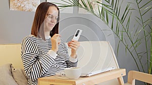 Smiling cheerful woman wearing striped shirt taking break at home workplace with closed laptop and coffee, reading message on