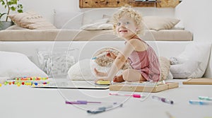 Smiling child, beautiful little girl with blue eyes and curly blonde hair, draws and writes cheerfully playing with coloured pens