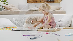 Smiling child, beautiful little girl with blue eyes and curly blonde hair, draws and writes cheerfully playing with coloured pens