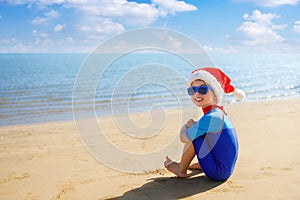 smiling child boy in santa hat, sunglasses and swimsuit sitting on the sand beach