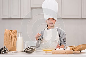 smiling child in chef hat whisking eggs in bowl at table