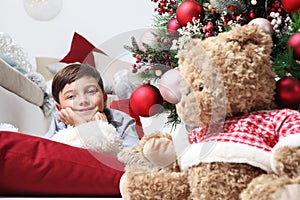 Smiling child and the Christmas tree decorated with balls, in the living room next to the teddy bear gift