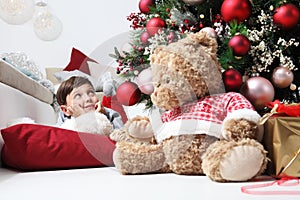 Smiling child and the Christmas tree decorated with balls, in the living room next to the teddy bear gift