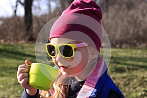 Smiling child in colorful clothes drinks tea from bright green reusable cup Spring nature background Eco-friendly picnic