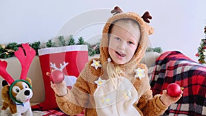 Smiling child in deer costume looking into the camera making funny face with red Christmas balls on her eyes.