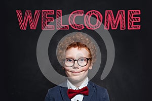 Smiling child with ginger hair portrait. Cute little boy in blue suit against blackboard with welcome inscription