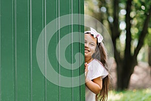 Smiling child girl looks out from behind a green wall in the summer outdoors