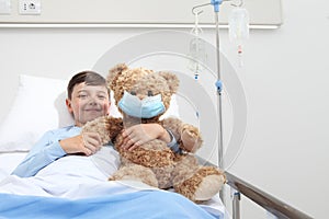 Smiling child in hospital bed with teddy bear wearing protective mask, corona virus covid 19 protection concept