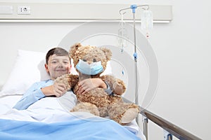 Smiling child in hospital bed with teddy bear wearing protective mask, corona virus covid 19 protection concept