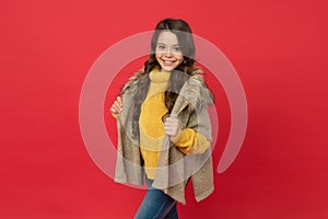Smiling child with long curly hair wear warm knitted sweater and fur waistcoat on red background, fashion and beauty