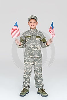 smiling child in military uniform with american flagpoles in hands looking at camera