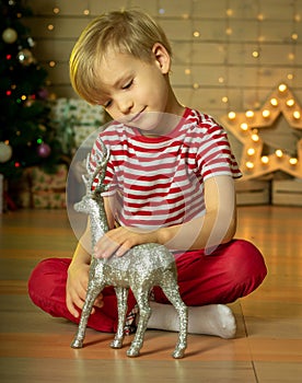 Smiling child in a red Santa Claus hat playing with new year silver deer with on the background of the christmas tree. New Year`s