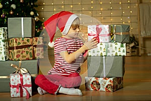 Smiling child in a red Santa Claus hat prepares a Christmas present on the background of a Christmas tree. New Year`s