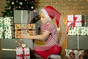Smiling child in a red Santa Claus hat prepares a Christmas present on the background of a Christmas tree. New Year`s