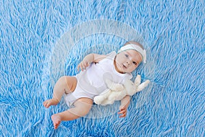 A smiling child in a white bodysuit lies on a blue fur background with a toy