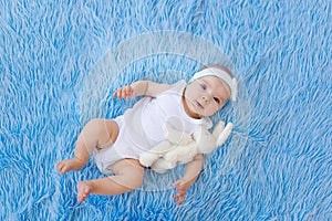 A smiling child in a white bodysuit lies on a blue fur background with a toy