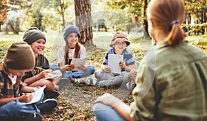 Smiling children and   female teacher sitting in circle on grass in forest and talking about nature