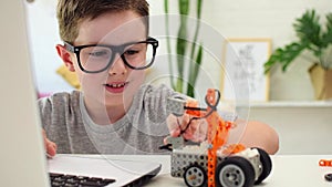 Smiling clever boy in eyeglasses programs a robot car using a laptop at home. The child is learning coding and