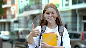 Smiling college girl with copybooks showing thumbs-up, satisfied with course