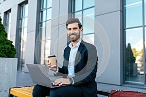 Smiling confident european millennial bearded businessman in suit work on laptop, drink coffee on bench