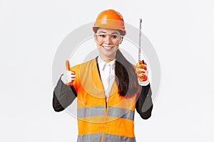 Smiling confident female asian construction engineer, architect in safety helmet, uniform, showing tape measure and