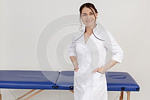 Smiling confident female doctor in medical overalls standing near massage table and looking at camera. Cosmetology spa