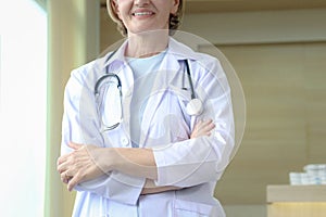 Smiling confident female doctor in white workwear with stethoscope, standing with arms crossed in clinic hospital. Professional