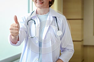 Smiling confident female doctor in white workwear with stethoscope, standing and giving thumb up in clinic hospital. Professional