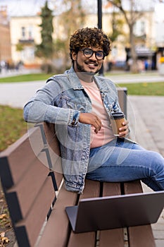 Smiling confident indian bearded businessman in suit work on laptop, drink coffee on bench near office building