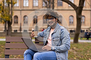 Smiling confident indian bearded businessman in suit work on laptop, drink coffee on bench near office building