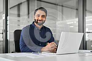 Smiling confident Indian business man sitting at work desk with laptop. Portrait