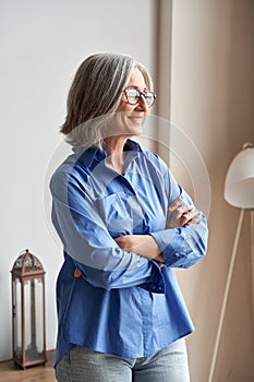 Smiling confident mature woman standing arms crossed indoors looking at window.