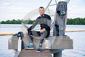 Smiling confident wakeboarder resting after training, sitting on pier with wakeboard