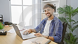 Smiling, confident young arab man joyously working on his laptop at the office, exuding positivity as the successful manager that